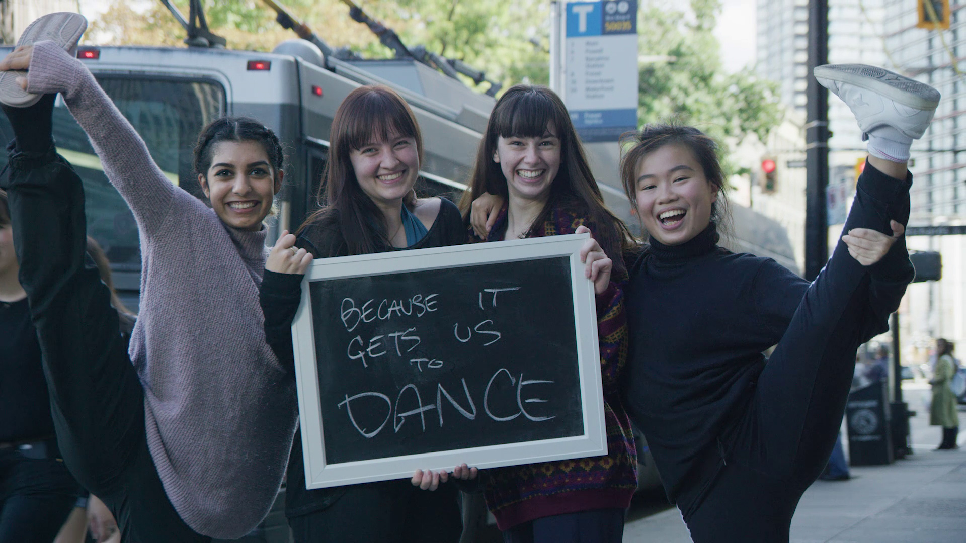 Four SFU School of Contemporary Arts students pose behind a chalk board that reads "Because it gets us to dance" 
