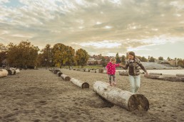A baby walks along a log at Kitsilano Beach with the help of their parent