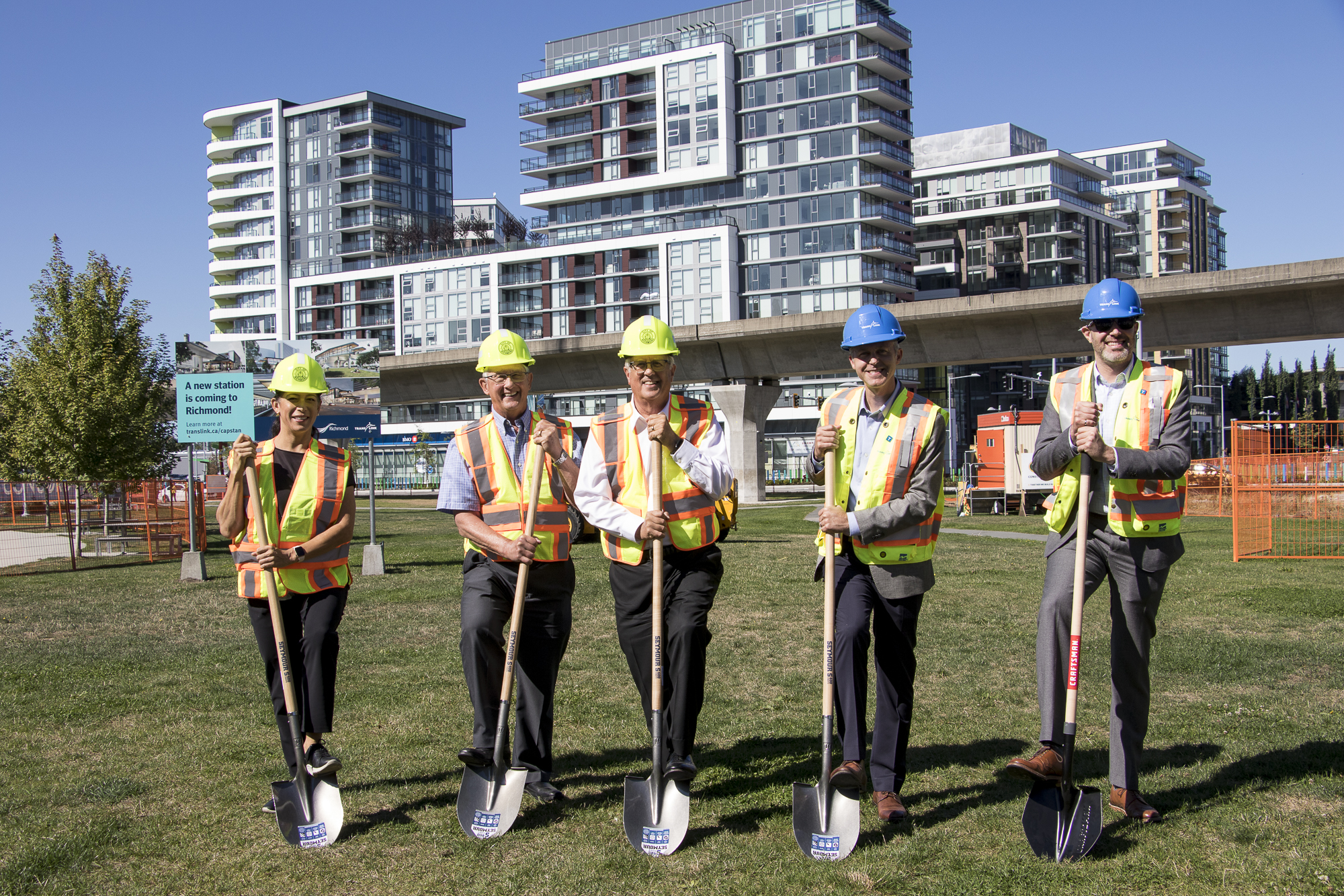 Various planner and politicians wear hard hats and high visibility vets while holding shovels on the grass where the new Capstan Station will be