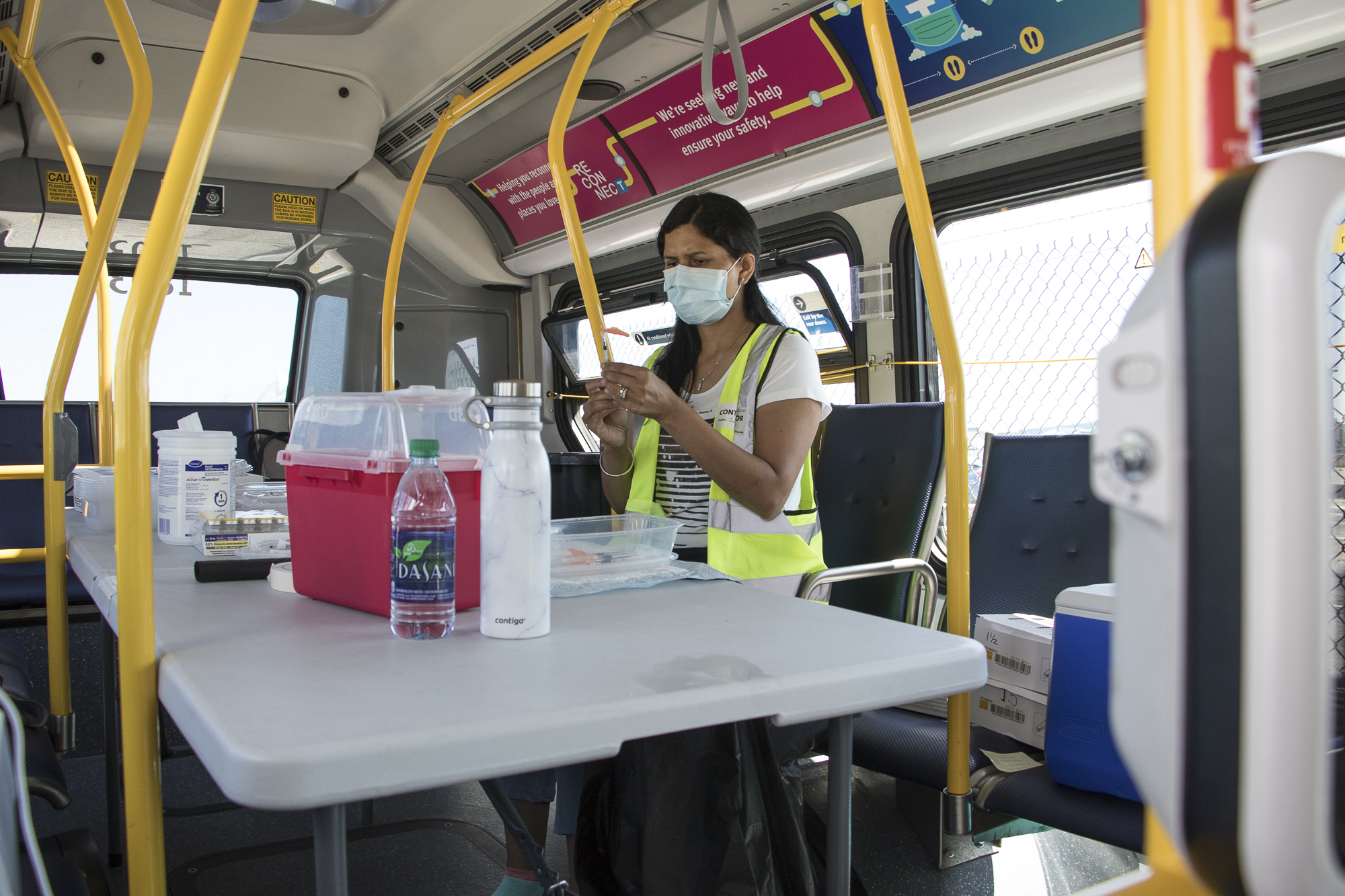 A Fraser Health nurse sits at the back of the bus preparing COVID-19 immunizations. She is masked and wearing a high visibility vest.