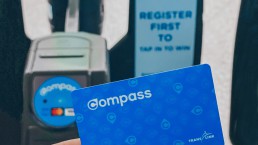 Holding up a Compass Card in front of fare gates with Tap In to Win decal