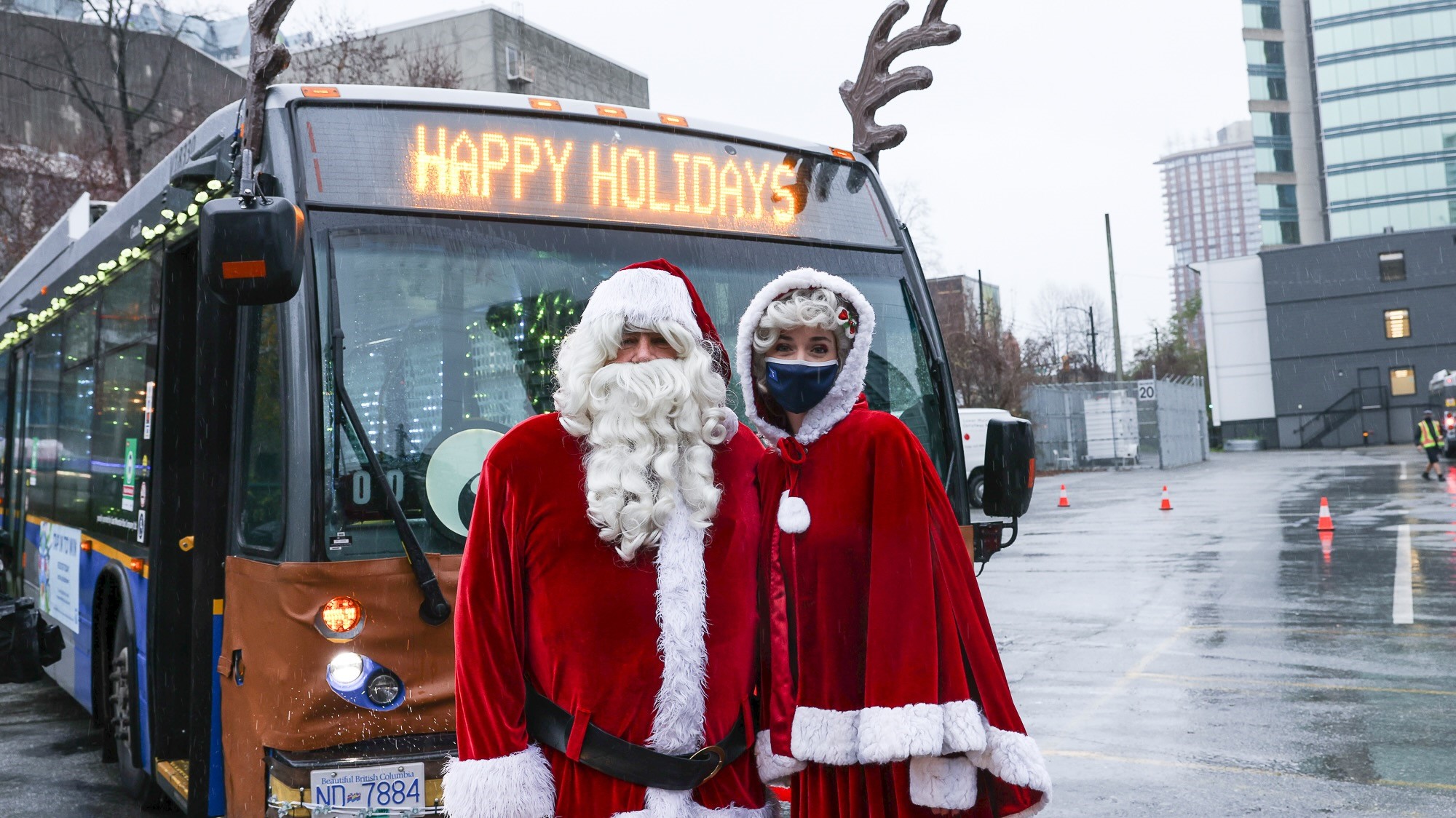 Santa Claus and Mrs Claus pose in front of a TransLink bus that is decorated as a Reindeer, with antlers and a destination sign that reads Happy Holidays