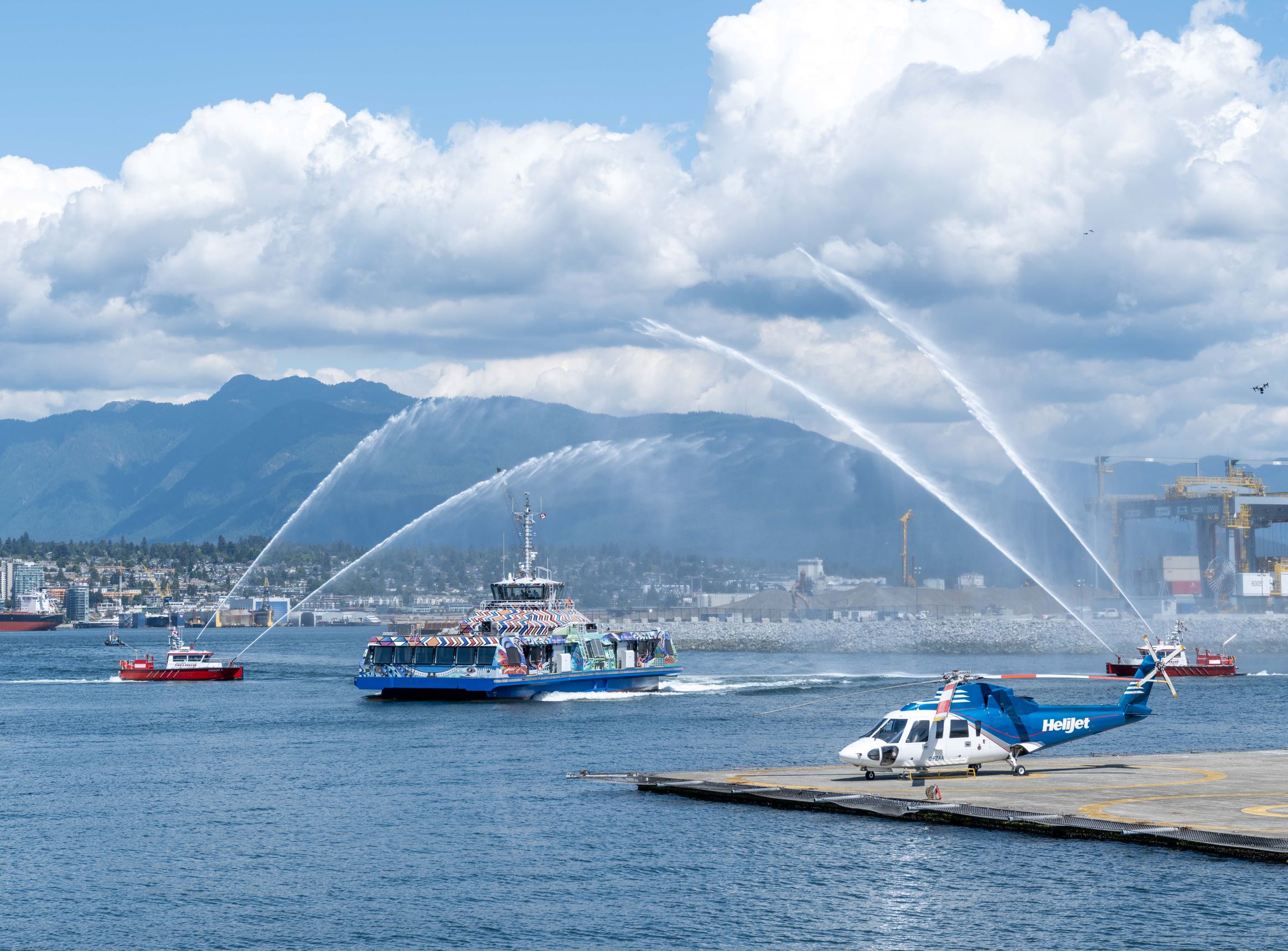 A SeaBus covered in Indigenous art is seen on the water between a water salute by two nearby Vancouver Fire Department boats.