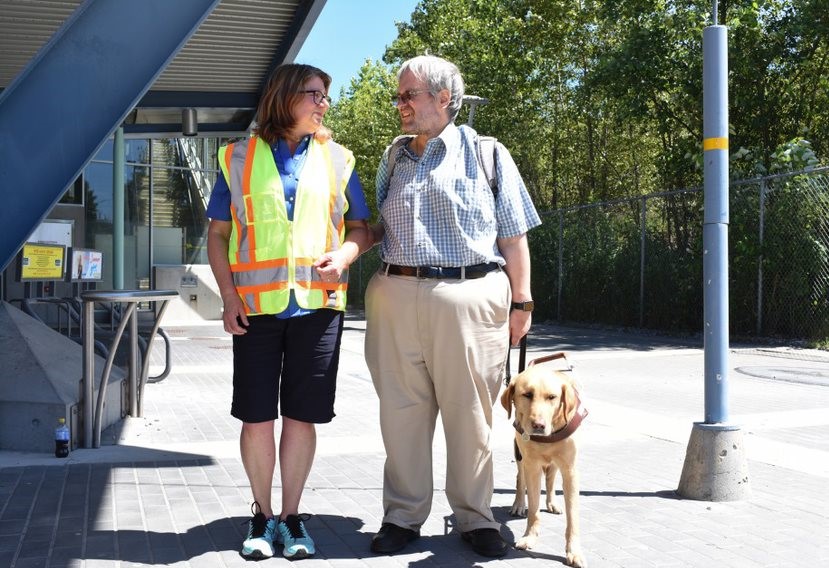 TransLink worker helping visually impaired customer with guide dog at transit station. 