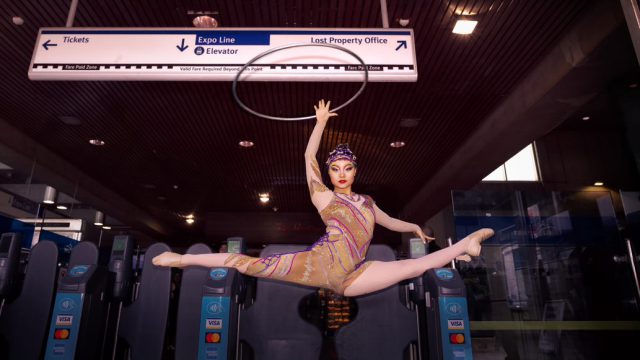Cirque du Soleil performer in costume holding hula hoop while doing the splits on top of fare gate at SkyTrain station.