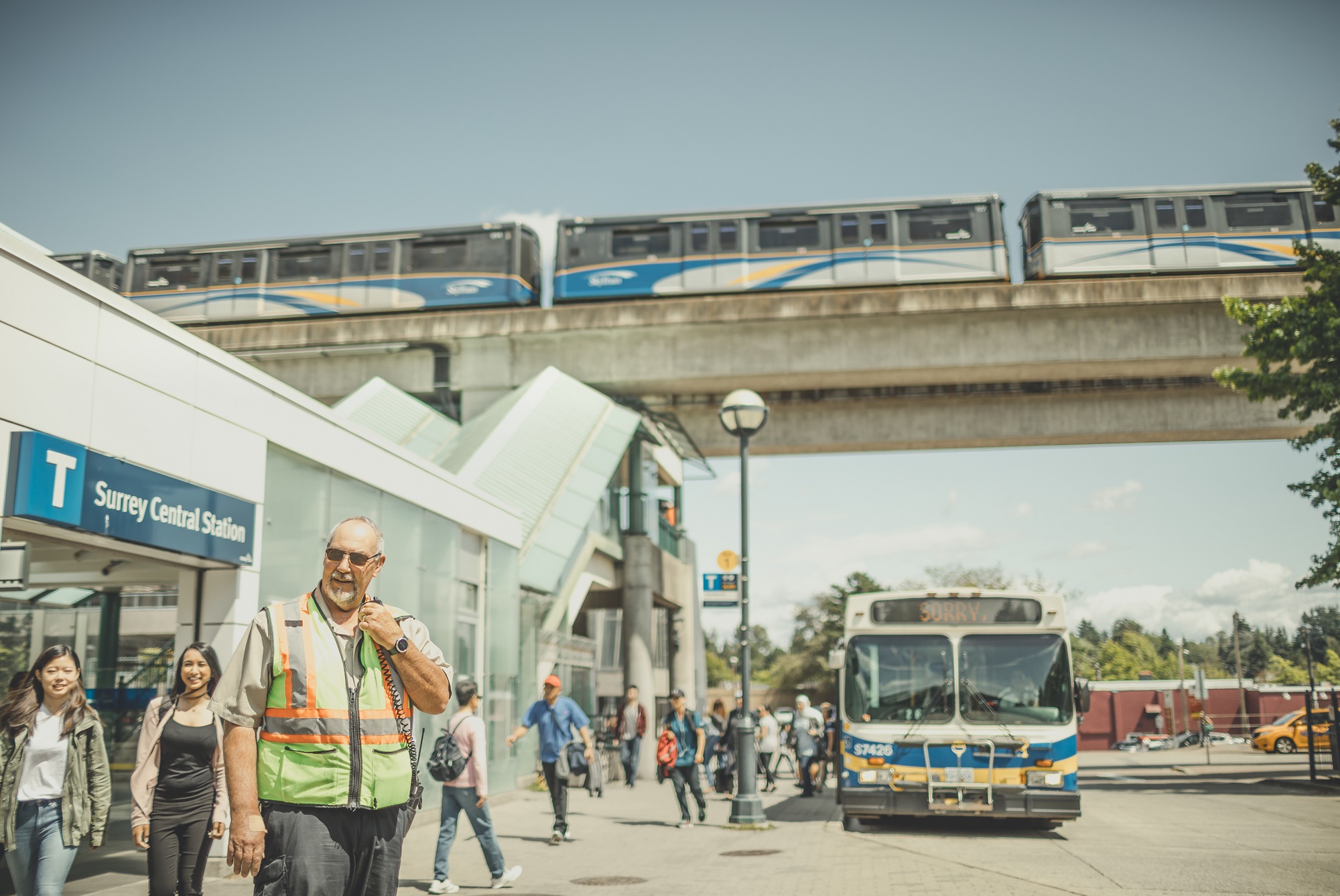 Man in a high vis vest stands in front of Surrey Central Station with a bus and SkyTrain in the background