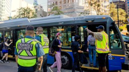 Transit employees wearing high-vis vests direct people aboard a bus that reads 