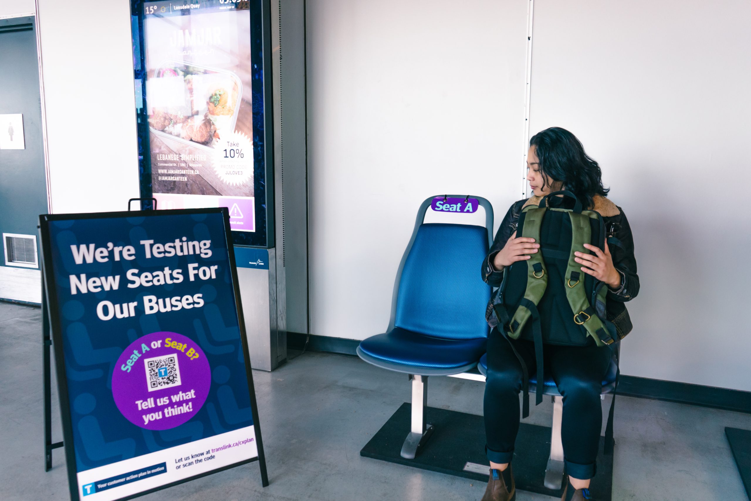 Bus seat testing site with information sandwich board on left and person sitting in bus seat on right, while looking at bus seat next to them on left.