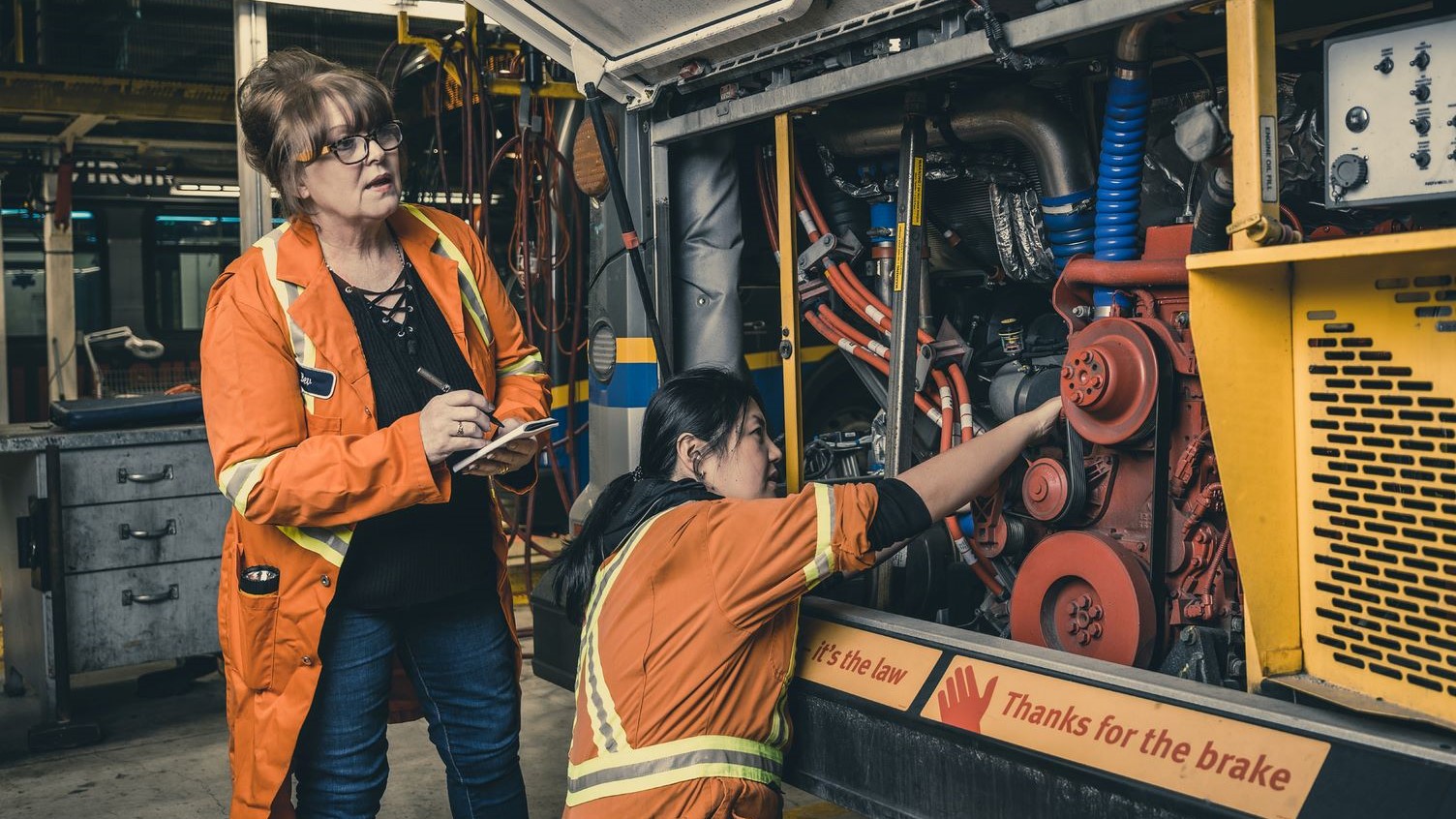A mechanic inspects the engine of a bus while another takes notes