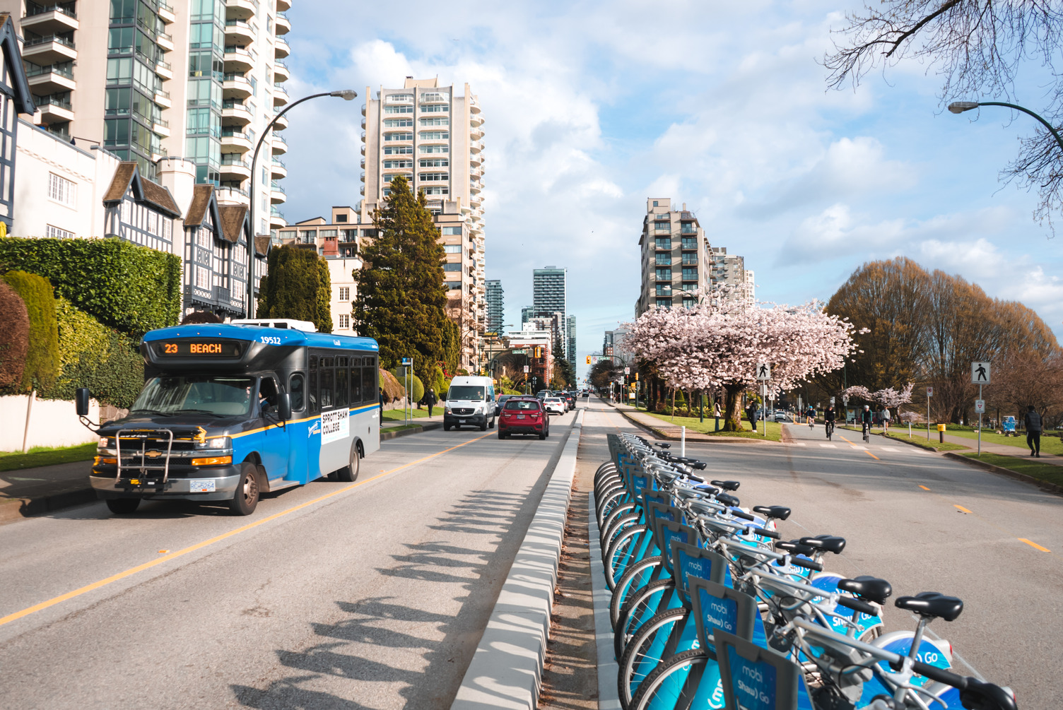 Beach bus driving through English Bay in spring past cherry blossoms and community bike racks. TransLink bus schedules are changing with Spring Service Changes.