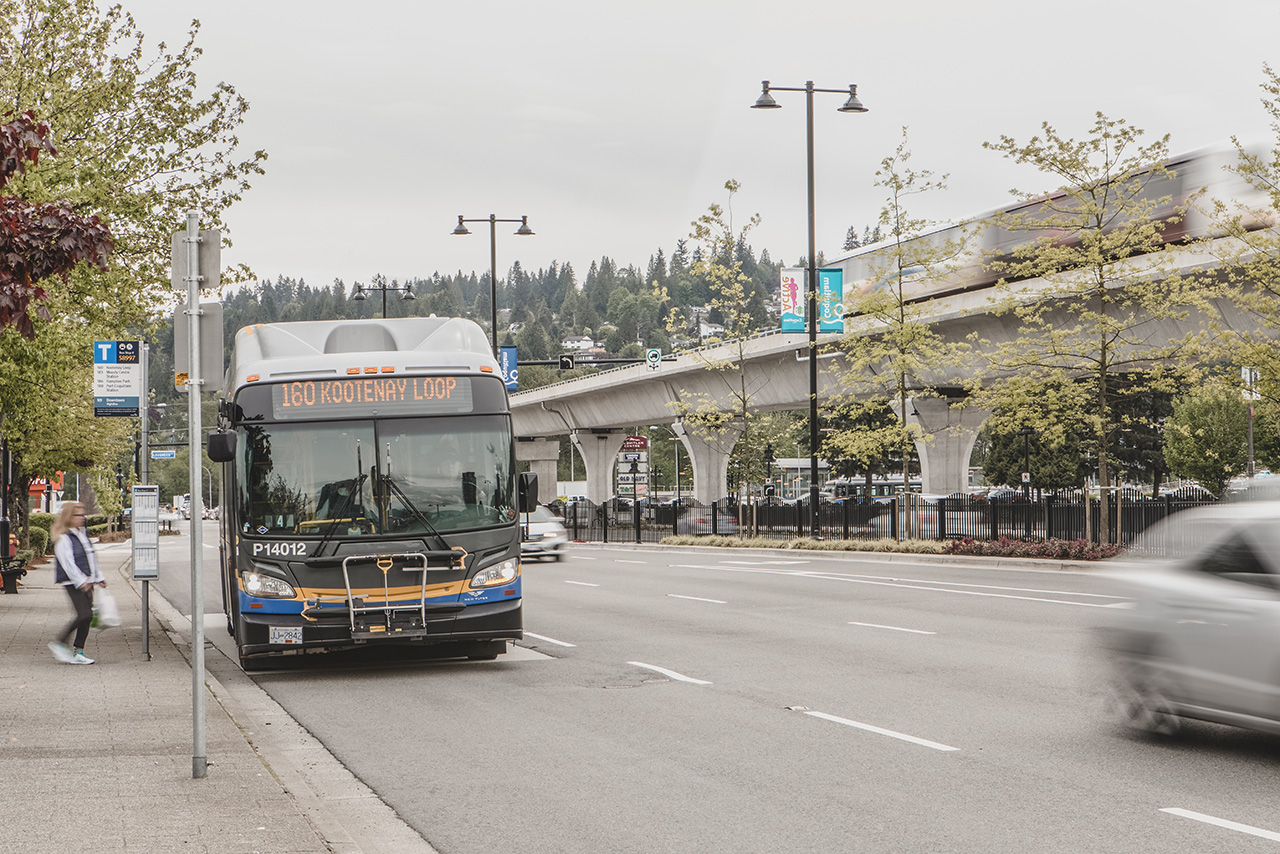 The 160 Kootenay Loop bus pulls into a bus stop as the SkyTrain passes by in the background