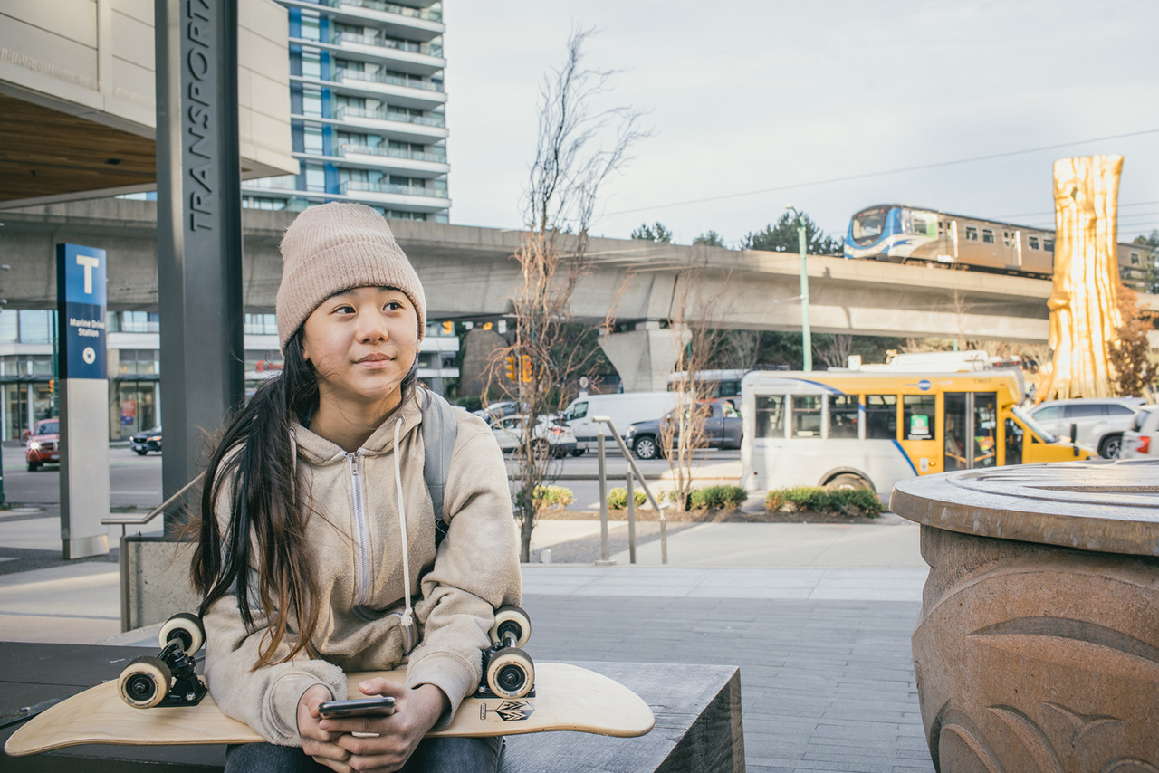 Young girl sitting near Marine Drive Station. SkyTrain and HandyDART bus pass by