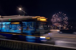 Bus and car passing crowd of people on bridge with fireworks in background