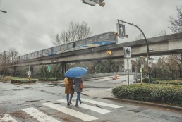 Two people crossing the road while the SkyTrain passes on the guideway. It's a rainy day.