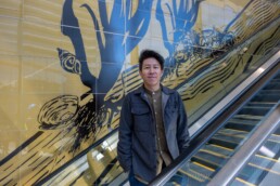 Arist Howie Tsui standing on an escalator with his art behind