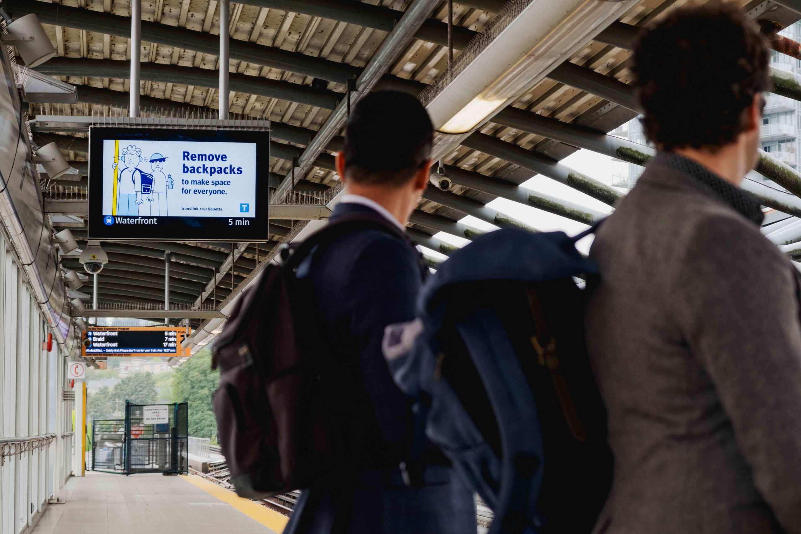 customers wait at the SkyTrain platform where a sign in the background displays a message about backpack transit etiquette