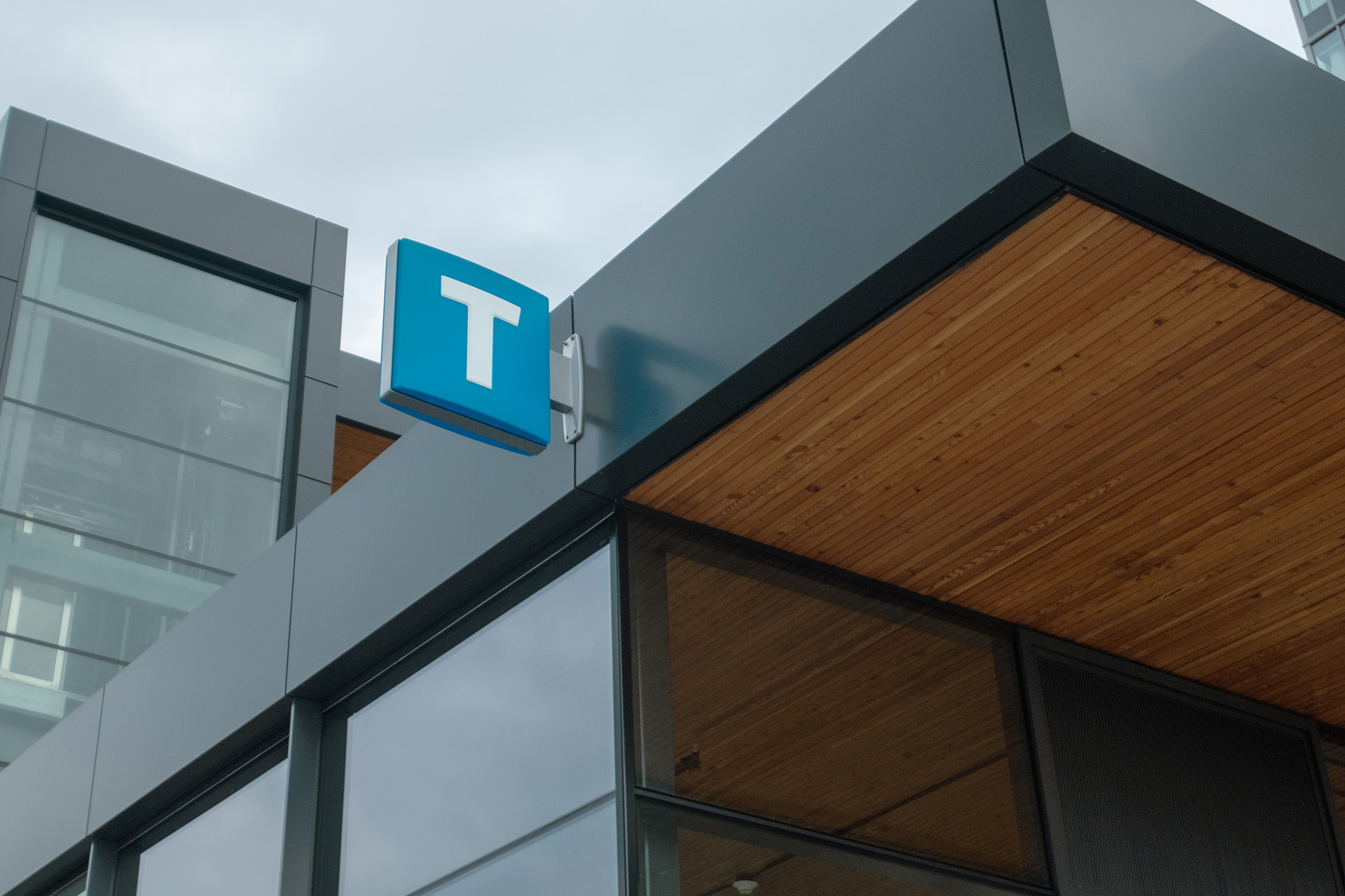 Metro Vancouver public transit sign showing a white capital letter 'T' on a blue square background, mounted on the exterior wall of a modern building. The 'T' indicates a transit station entrance.