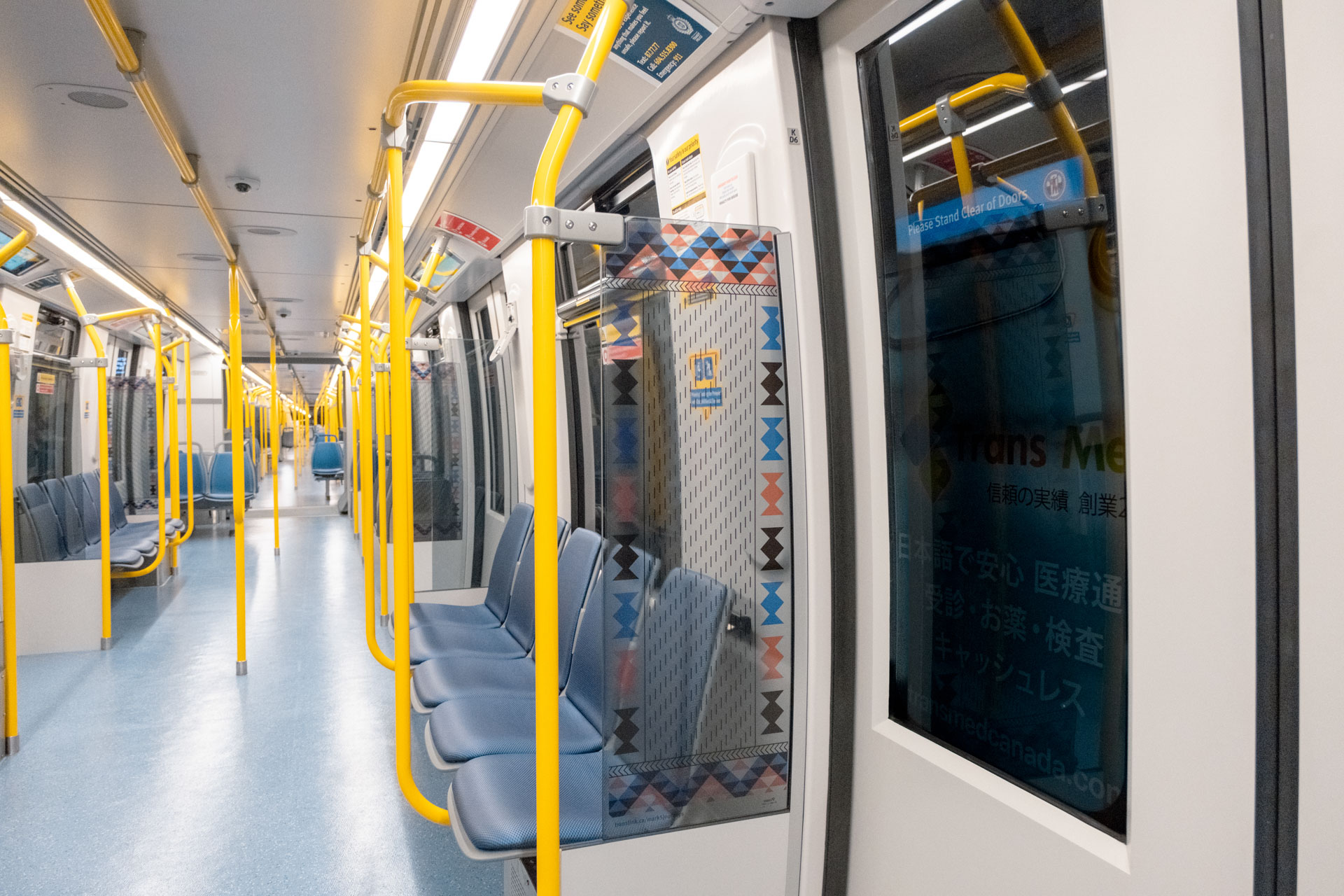 Interior of the new Mark V SkyTrain featuring blue side-facing seats, yellow poles, and a patterned artwork titled 'Path of the Ancestors, Path of Our Future' by Yususult (Rosemarie Williams) displayed on the windscreen, with geometric designs in blue, black, and red.