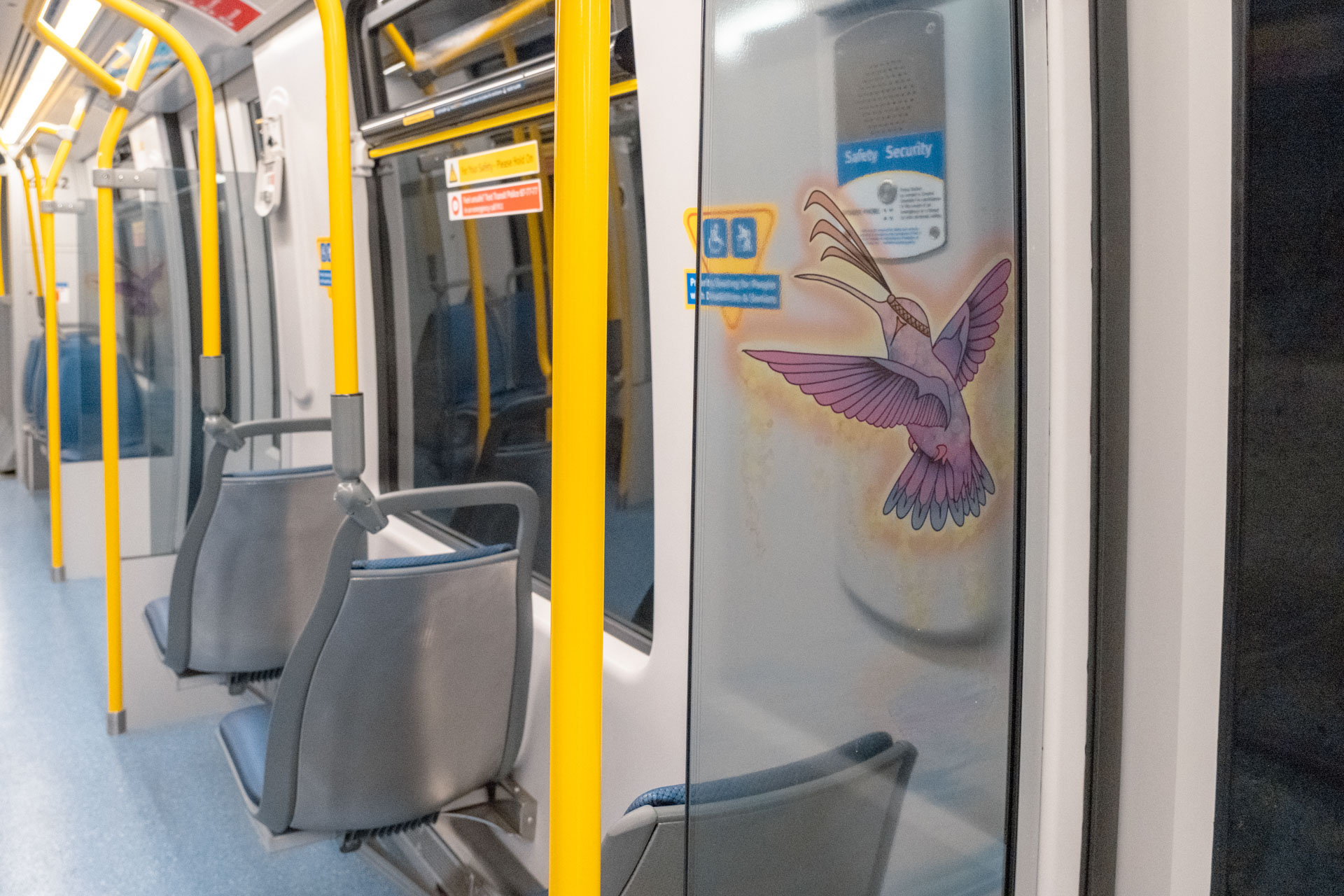 Interior of the new Mark V SkyTrain featuring grey and blue seats, yellow poles, and the artwork 'Hummingbird's Dance' by Rain Pierre on the right side, depicting a vibrantly coloured hummingbird in flight with detailed feathers.