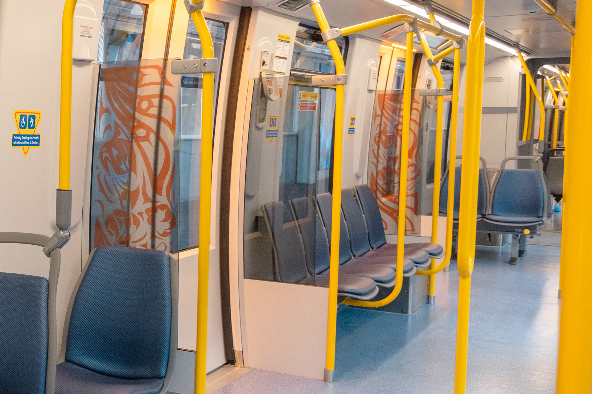 Interior of the new Mark V SkyTrain featuring blue side-facing seats, yellow poles, and the artwork 'Swooping Eagle' by Darryl Blyth displayed on the windscreen, depicting a bold, abstract eagle in flight using striking single-colour shapes and contrasting positive and negative space.