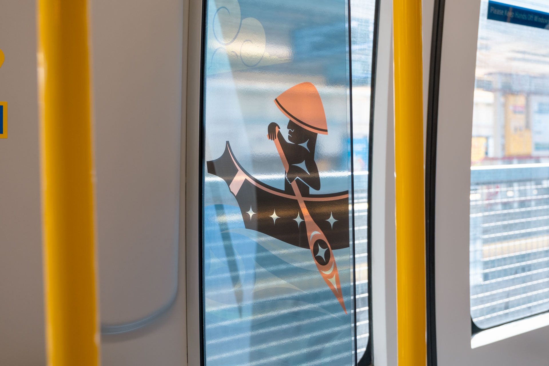 Interior of the new Mark V SkyTrain featuring the artwork 'Journey by Canoe: Paddling Through Salish Seas' by Atheana Picha on the windscreen, showing a stylised figure in a traditional hat paddling a decorated canoe through abstract blue-toned waves, with brown, black, and orange accents.