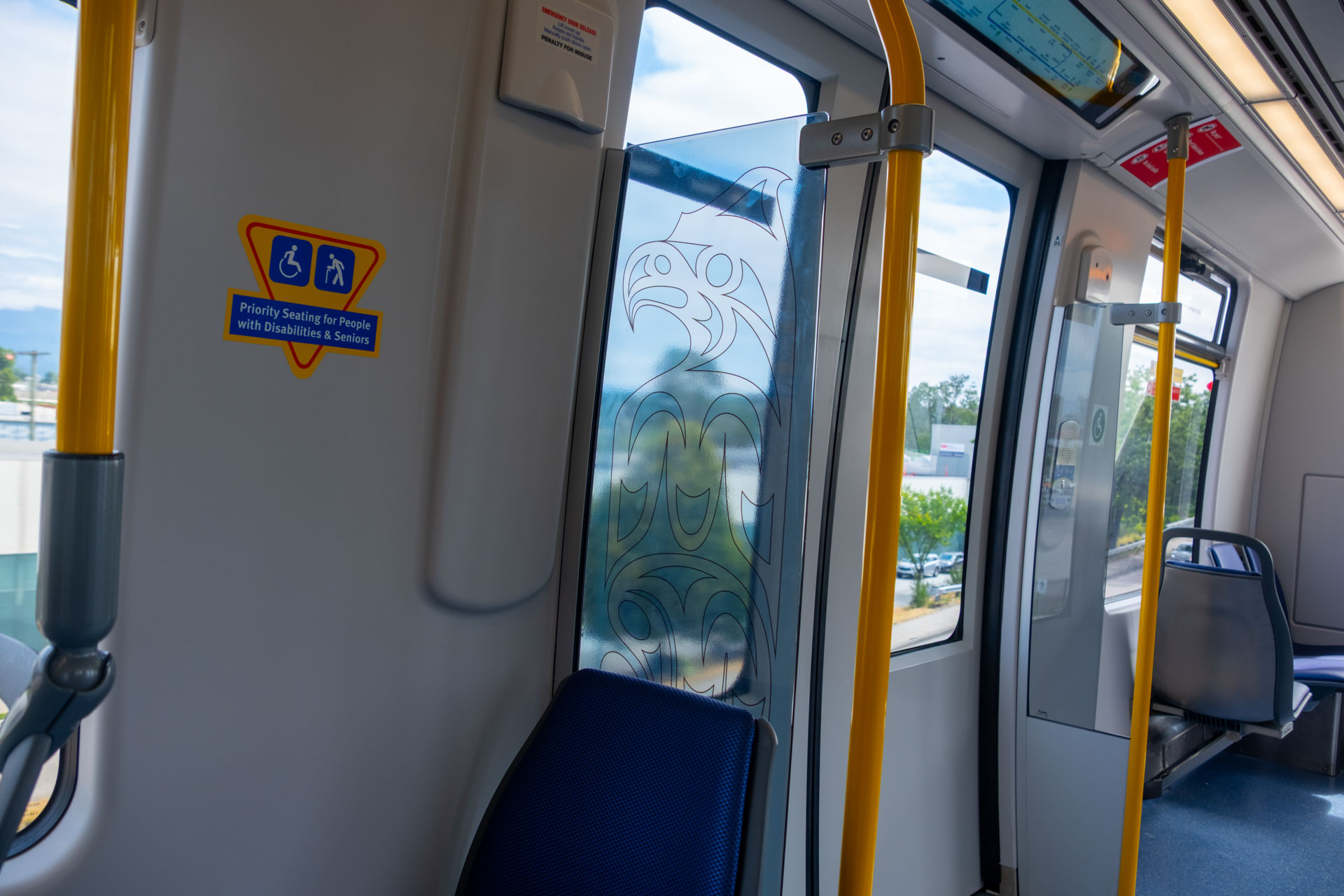 Interior of the new Mark V SkyTrain featuring blue seats, yellow poles, and the artwork 'Clearing the Path Forward' by Brandon Gabriel displayed on the windscreen, depicting a thunderbird in an Indigenous art style next to the priority seating area.