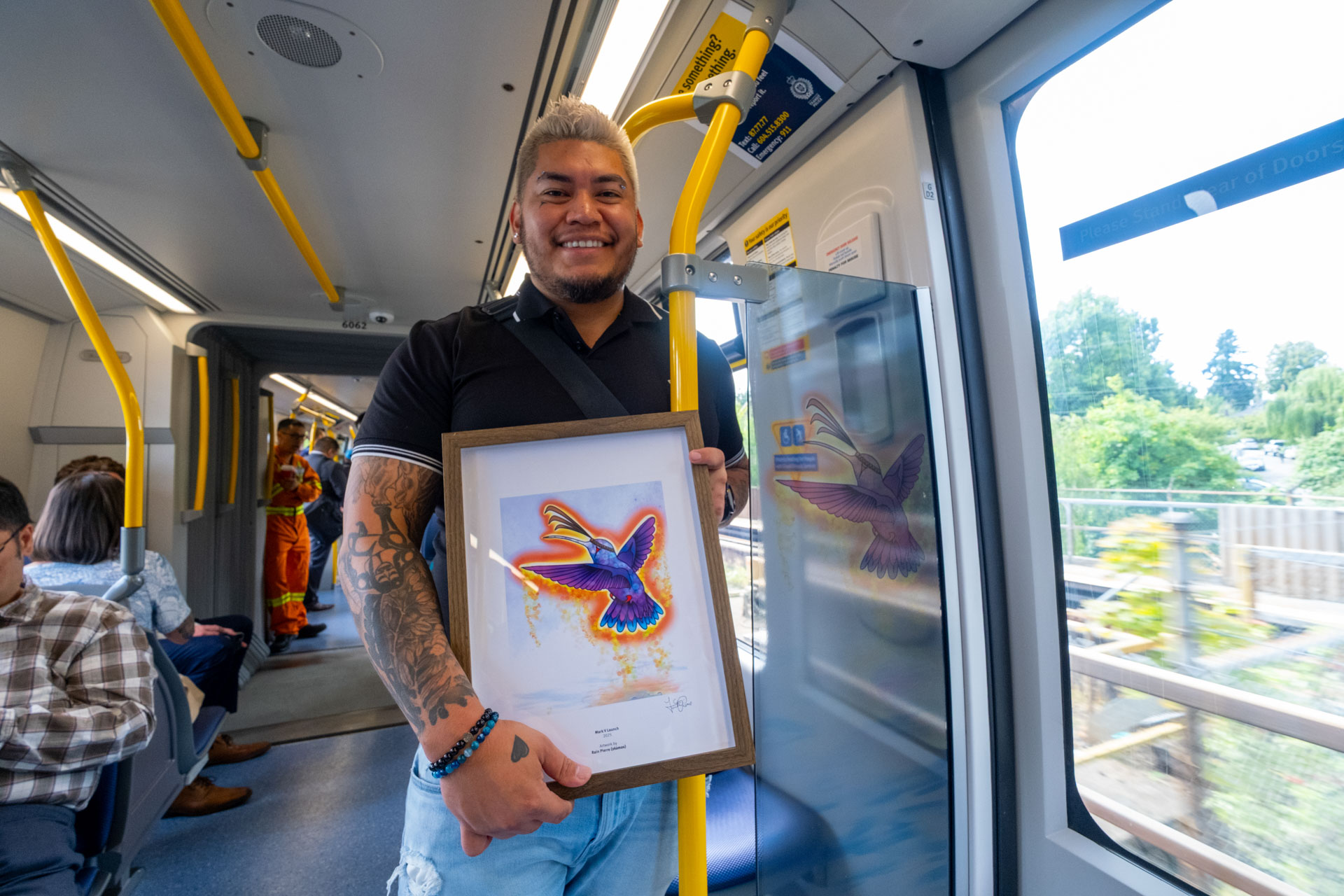 Inside the new Mark V SkyTrain, artist Rain Pierre stands holding a framed picture of his artwork 'Hummingbird's Dance,' with seated passengers in the background and motion blur visible through the windows, capturing the moment alongside his art on the train.