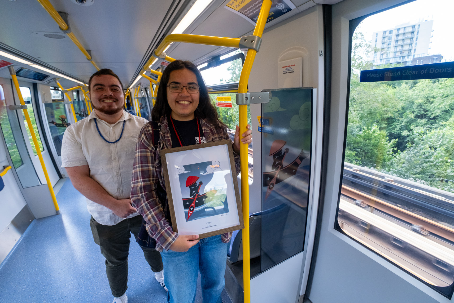 Inside the new Mark V SkyTrain, artist Atheana Picha stands on the right holding a framed picture of her artwork, with yellow handrails and large windows in the background. The image captures her alongside her art, 'Journey by Canoe: Paddling Through Salish Seas,' displayed on the train.
