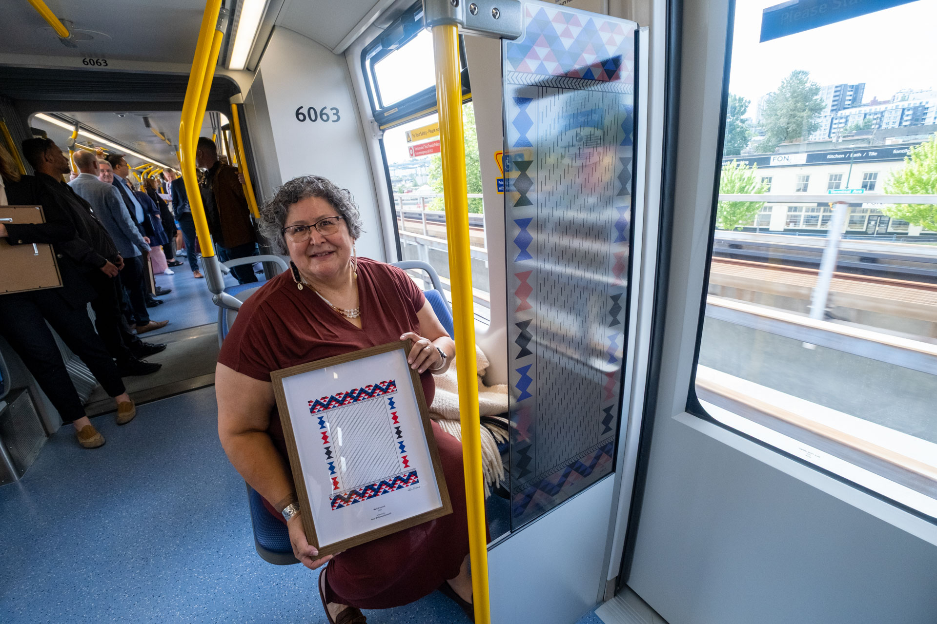 Inside the new Mark V SkyTrain, artist Rosemarie Williams sits near the windscreen where her artwork 'Path of the Ancestors, Path of Our Future' is displayed, holding a framed version of the piece, with other passengers in the background and greenery visible through the windows