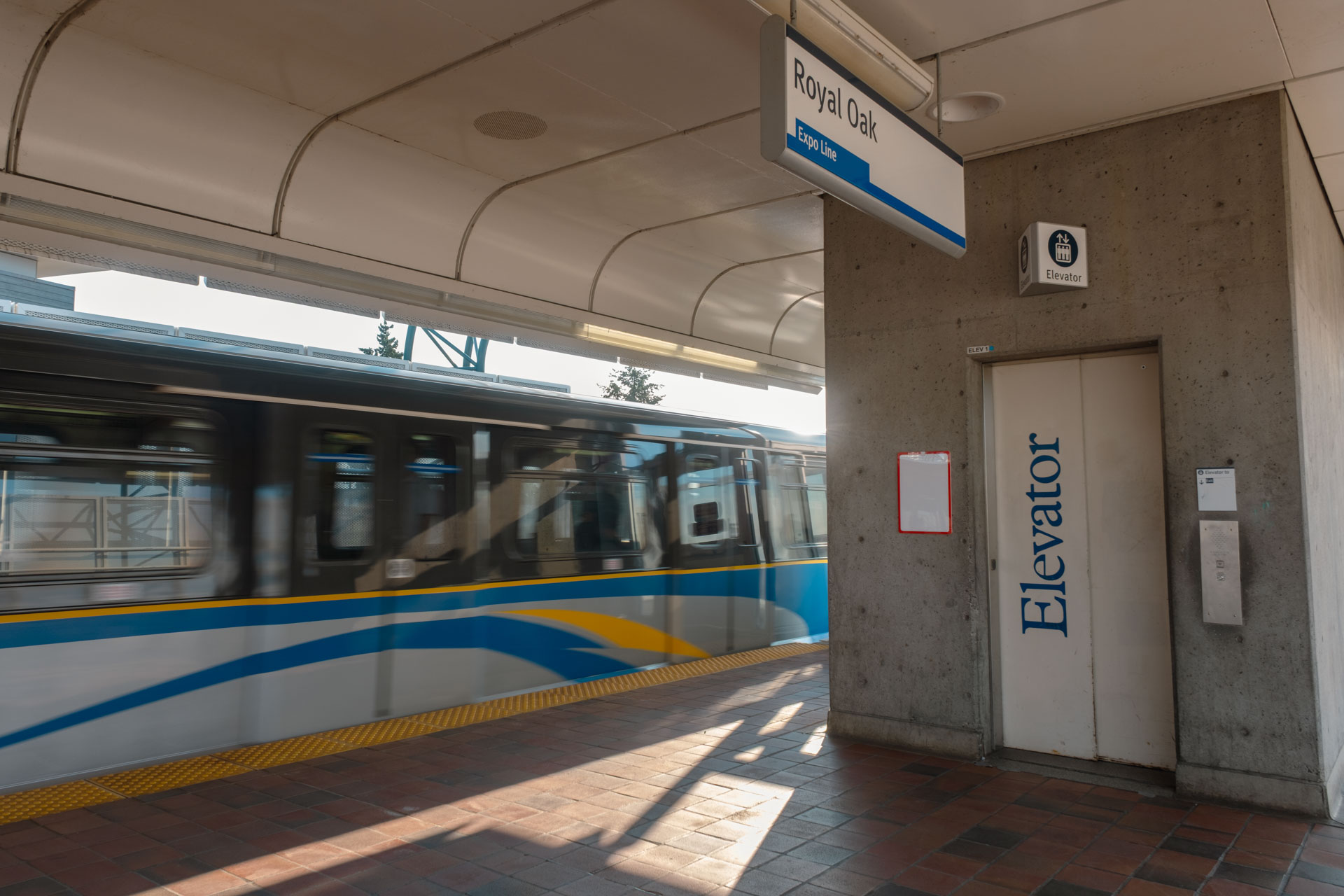 Royal Oak SkyTrain station platform with a moving train on the left featuring blue and yellow stripes. A sign above reads 'Royal Oak' and 'Expo Line'. On the right, an elevator door marked 'Elevator' includes accessibility signage and an intercom system. Sunlight casts shadows across the tiled platform and concrete walls.