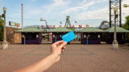 A hand holds up a blue Compass Card, with gates to Playland in the background