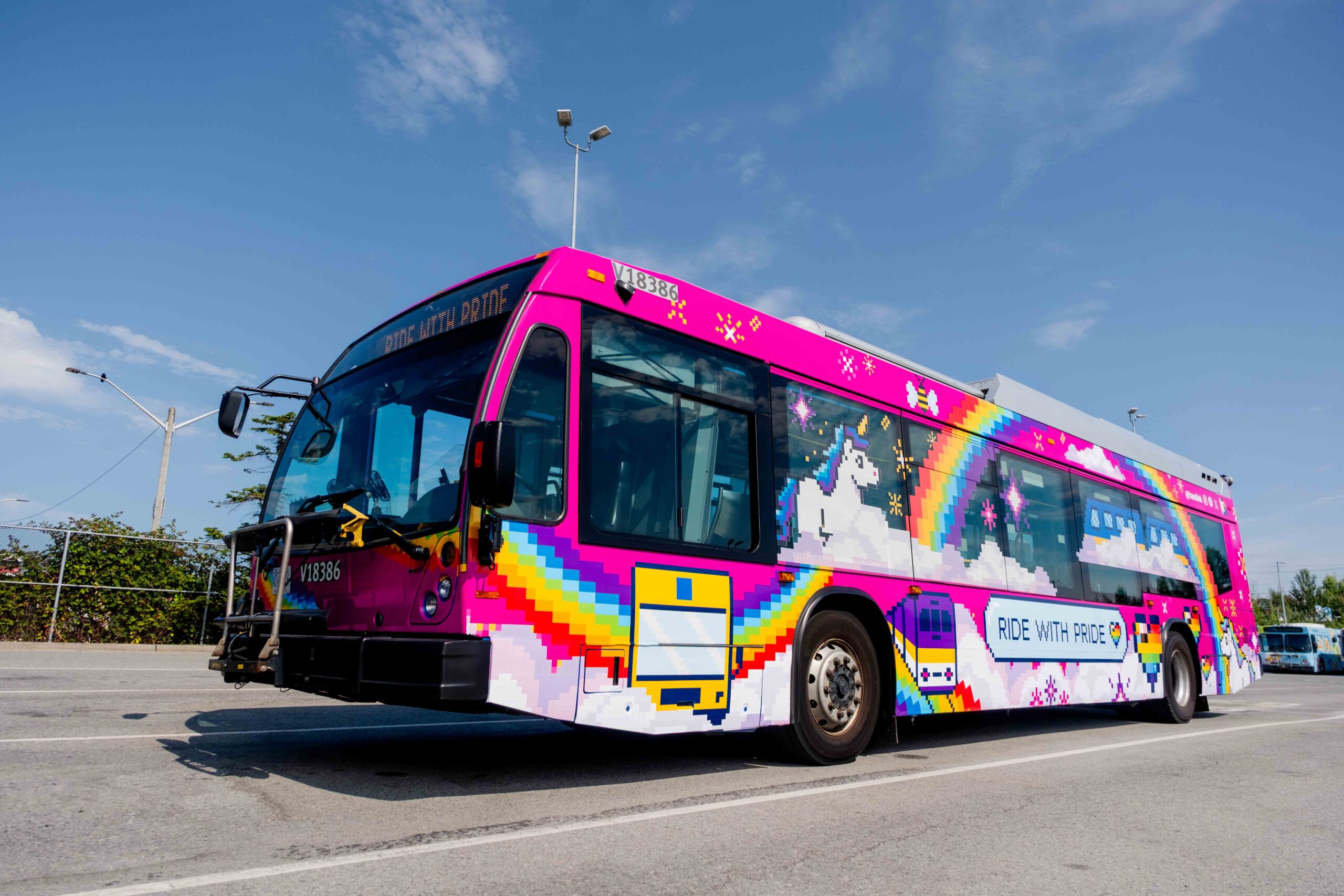 A colorful bus with pixel art of rainbows, unicorns, clouds, hearts, and stars on a pink background. The side of the bus displays the phrase 'RIDE WITH PRIDE' in blue letters on a white cloud.
