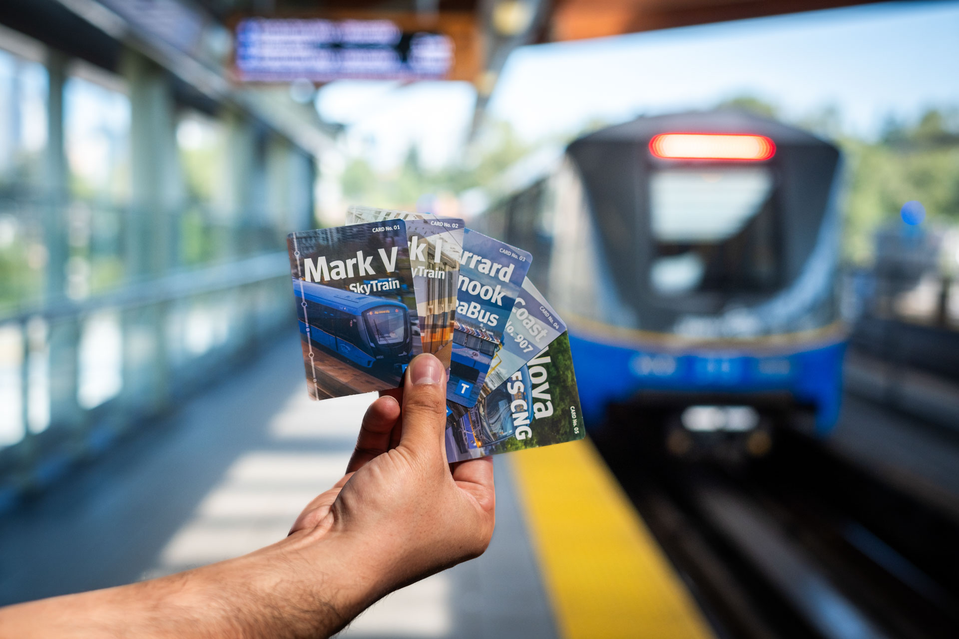 A hand holding the five TransLink trading cards on a SkyTrain train station platform with the train approaching in the background. The station features glass walls and a yellow safety line along the platform edge.