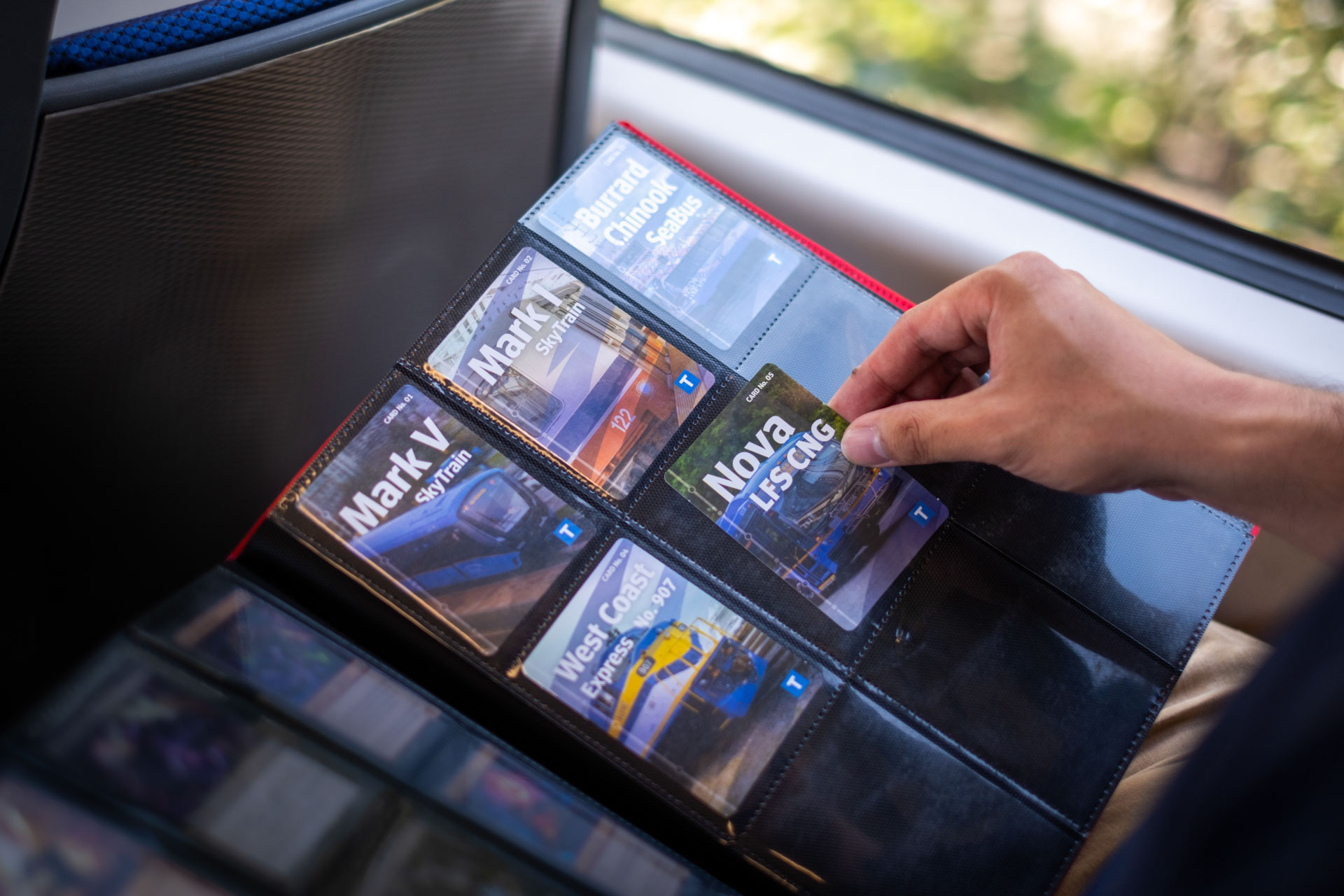 A person seated on the SkyTrain, holding a binder with plastic sleeves containing the TransLink trading cards.