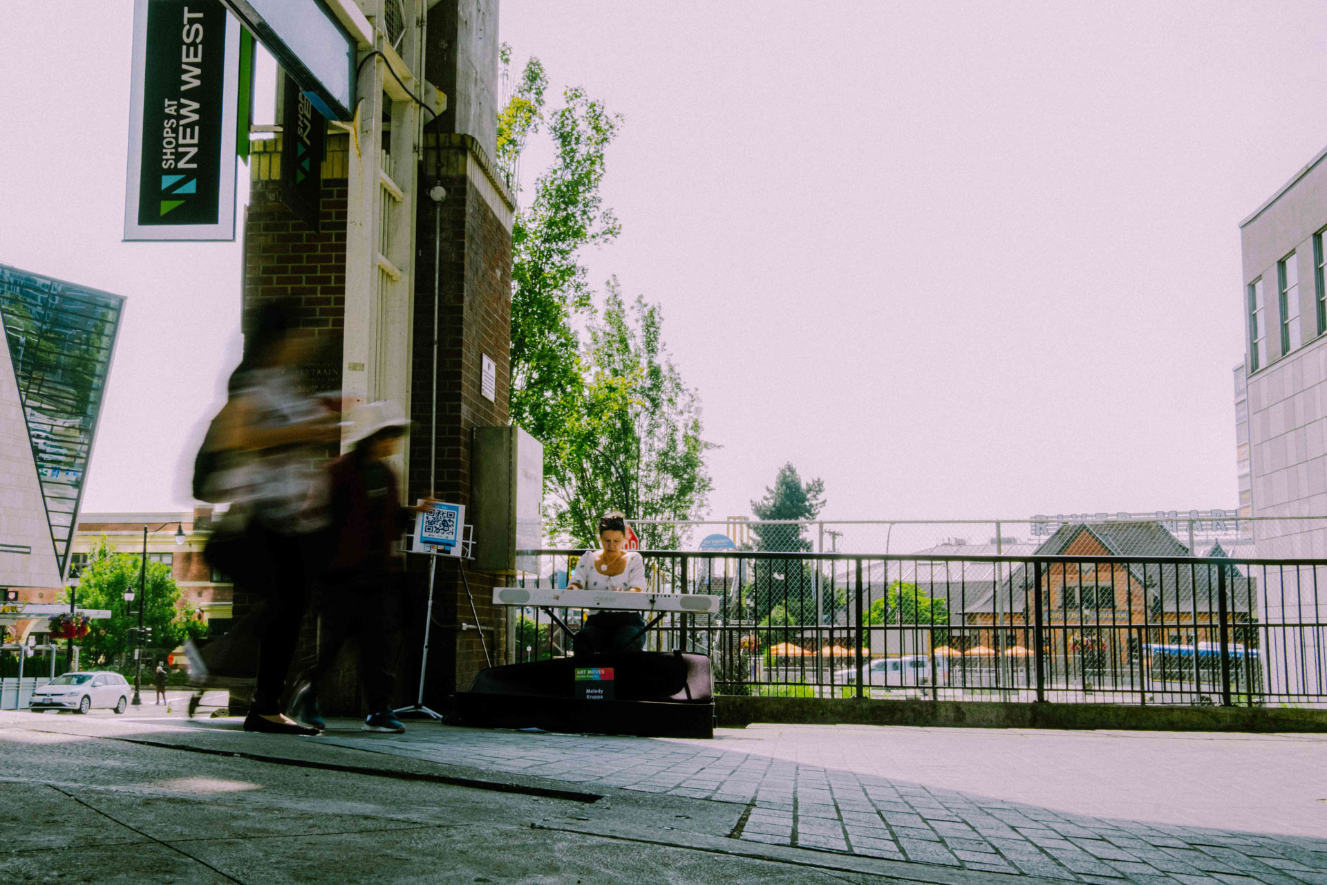 A musician playing a keyboard outside the Shops at New West entrance as pedestrians walk past on the sidewalk.