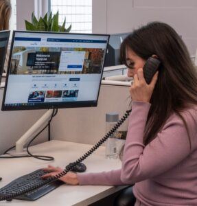 Office staff member speaking on the phone at a workstation with the TransLink website open on the computer screen.