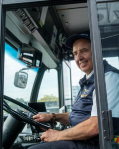 Transit operator seated at the driver’s seat of a Coast Mountain Bus Company vehicle, holding the steering wheel and preparing for service.