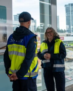 Two SkyTrain staff members in high‑visibility vests talking inside a station near large windows.