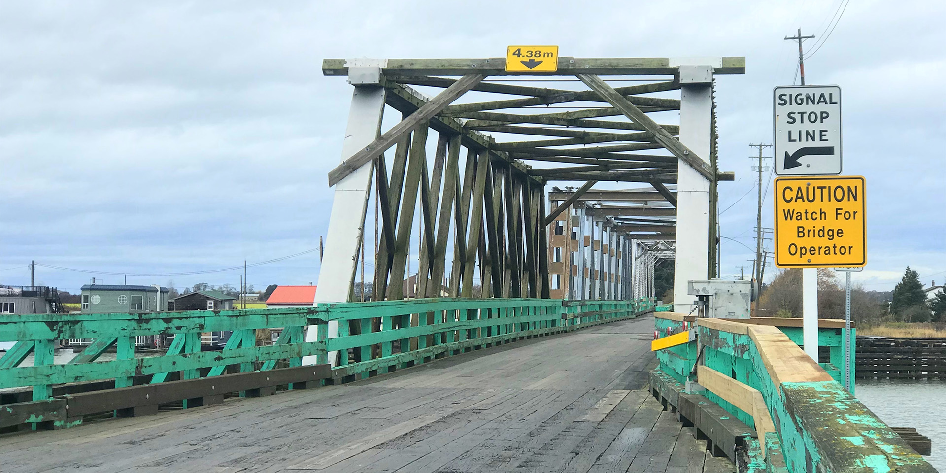 View of the Westham Island Bridge in Delta, a single‑lane wooden truss bridge with green railings and warning signs at the entrance under an overcast sky.