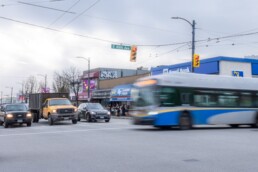 TransLink’s 49 bus traveling eastbound through the intersection of Fraser Street and East 49th Avenue, passing in front of shops and other vehicles on a cloudy day.