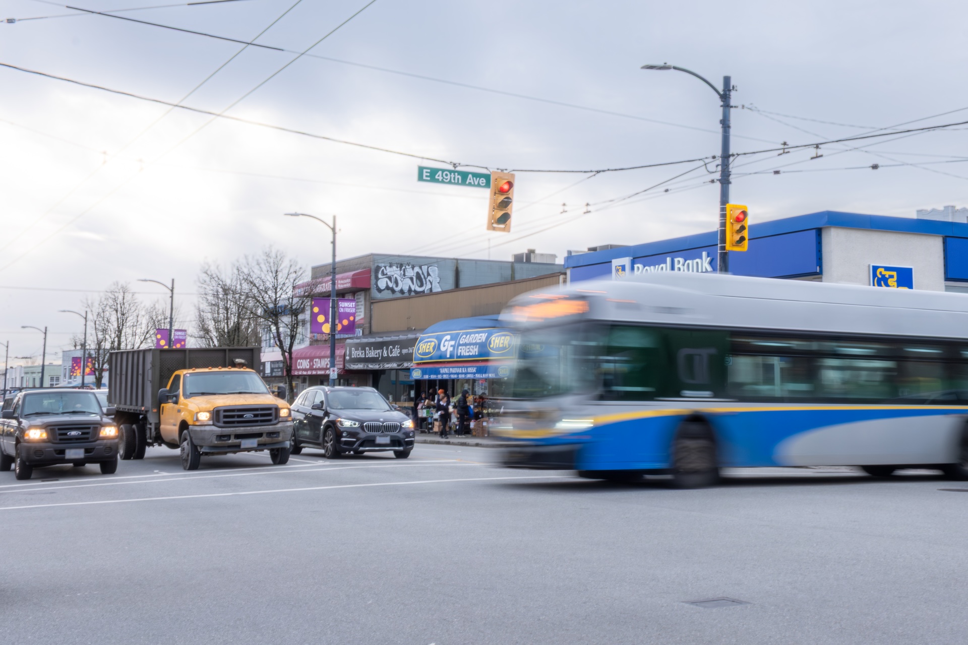 TransLink’s 49 bus traveling eastbound through the intersection of Fraser Street and East 49th Avenue, passing in front of shops and other vehicles on a cloudy day.