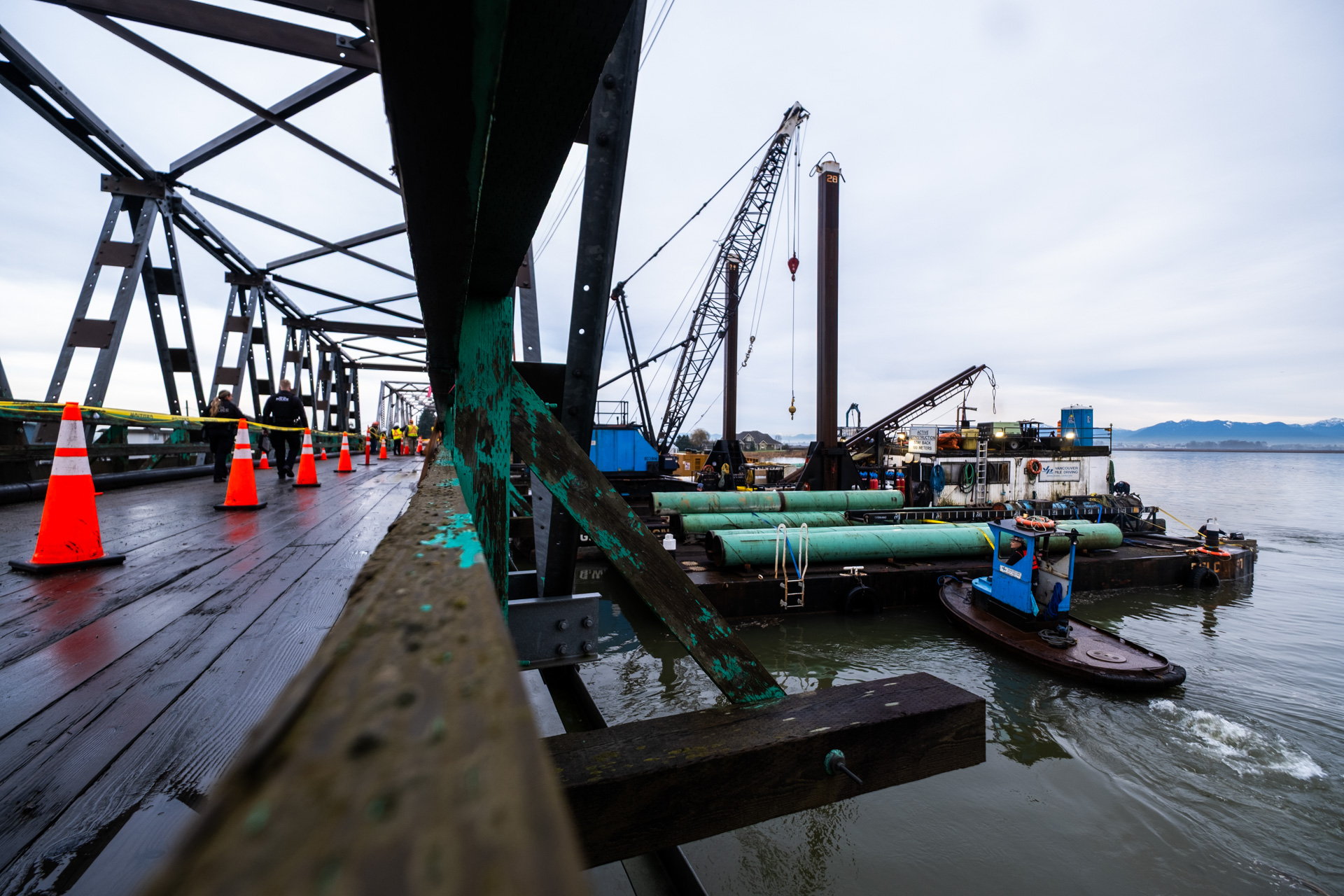 Close-up view from the deck of the Westham Island Bridge showing traffic cones and nearby construction barges with cranes working on repairs.