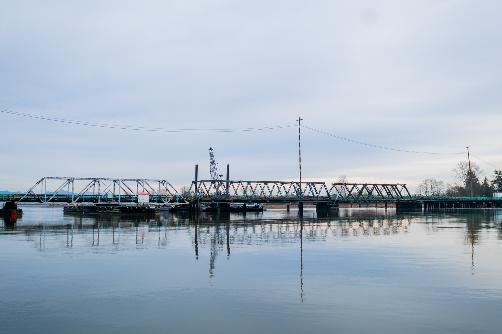 The Westham Island Bridge spanning across calm water under an overcast sky, with construction equipment visible in the distance.
