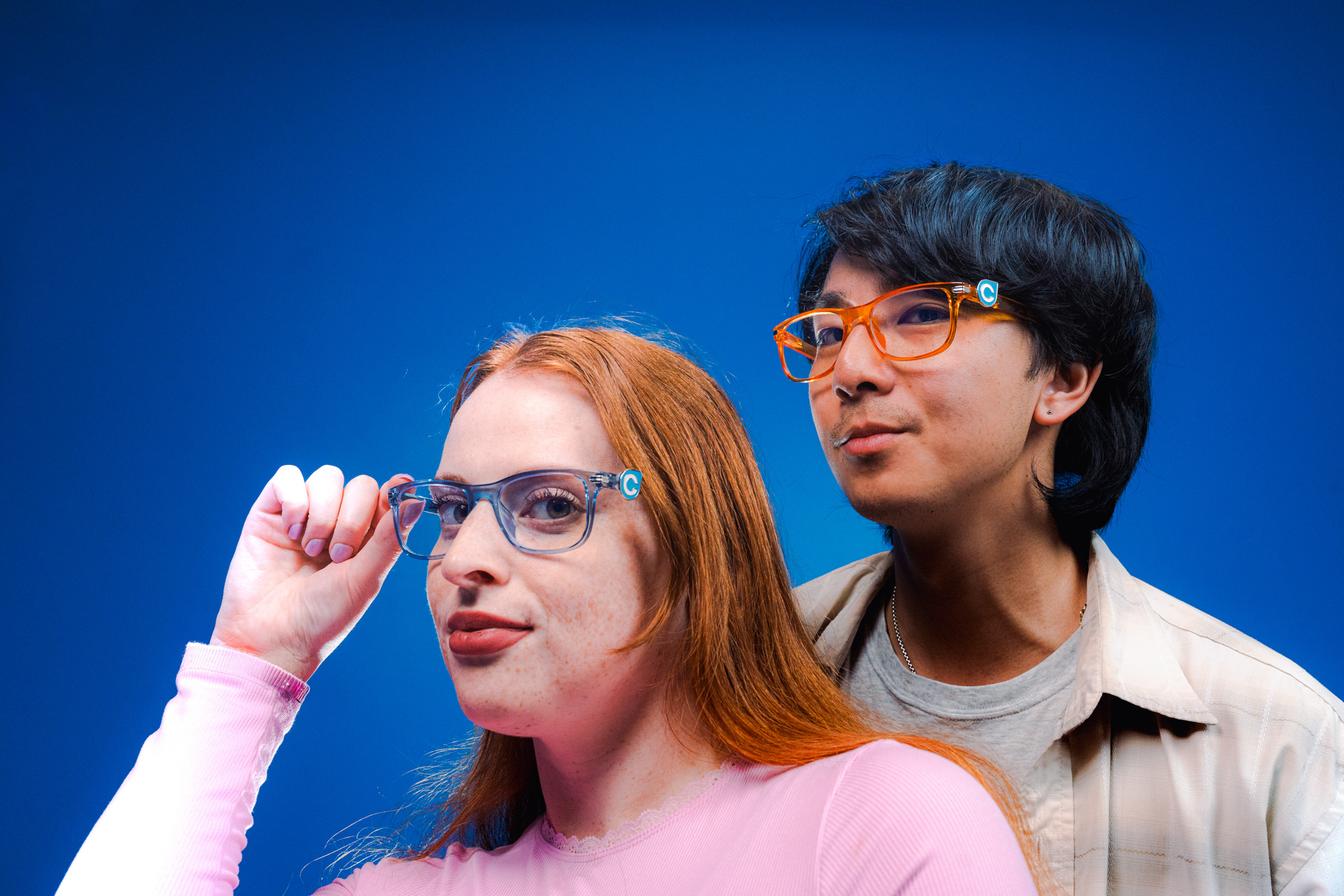 Two people wearing Compass Glasses pose against a blue background, one adjusting their clear frames and the other wearing orange frames.