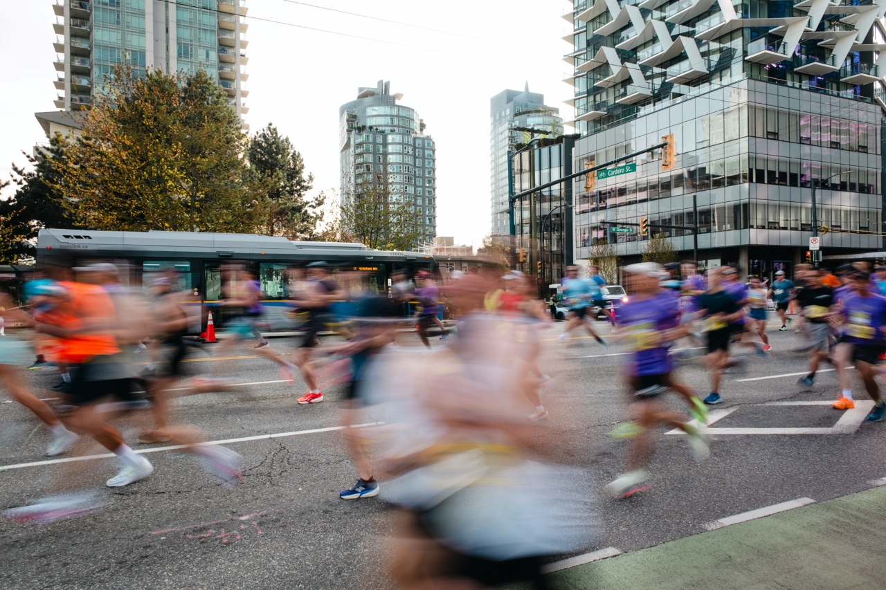 Motion-blurred runners race through a downtown Vancouver intersection during a marathon as a TransLink bus passes in the background, framed by high-rise buildings and early morning light.