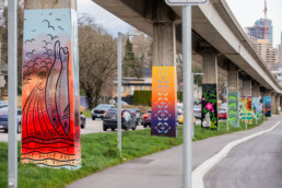 Colourful Indigenous artwork panels mounted on SkyTrain guideway pillars along Stewardson Way in New Westminster, beside the BC Parkway walking and cycling path.
