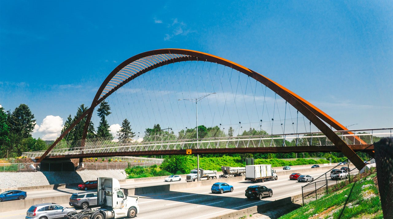 A pedestrian and cycling bridge with a large rust-coloured arch spans Highway 1 in Metro Vancouver, with vehicles travelling below under a bright blue sky.